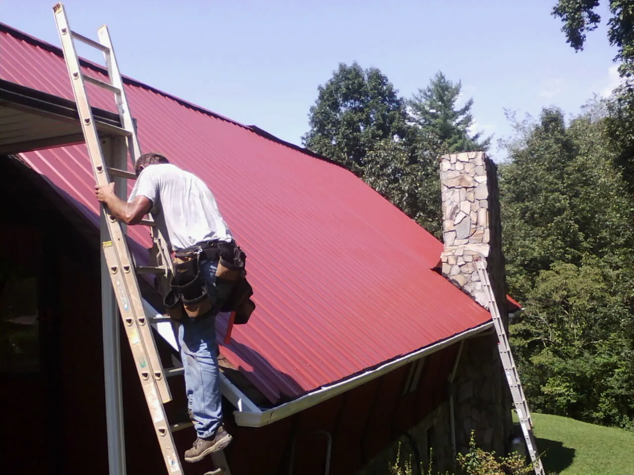 Roofer on a ladder beside a red metal roof and stone chimney — Soffit & Fascia Repair by Roofing Brothers in Harbor City